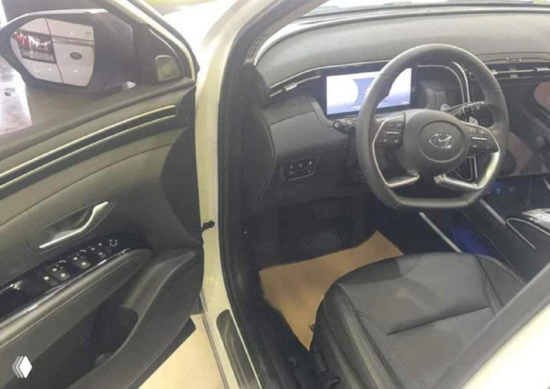 Руль, панель приборов и водительское место Hyundai Tucson: чёрные отделки, органы управления на двери и вид переднего сиденья в дилерском помещении.