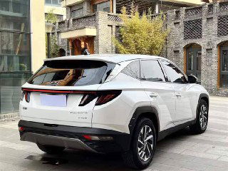 Задняя проекция белого Hyundai Tucson 2022 года: открытая кормовая часть, фон городских зданий и дизайн задних фонарей хорошо видны.