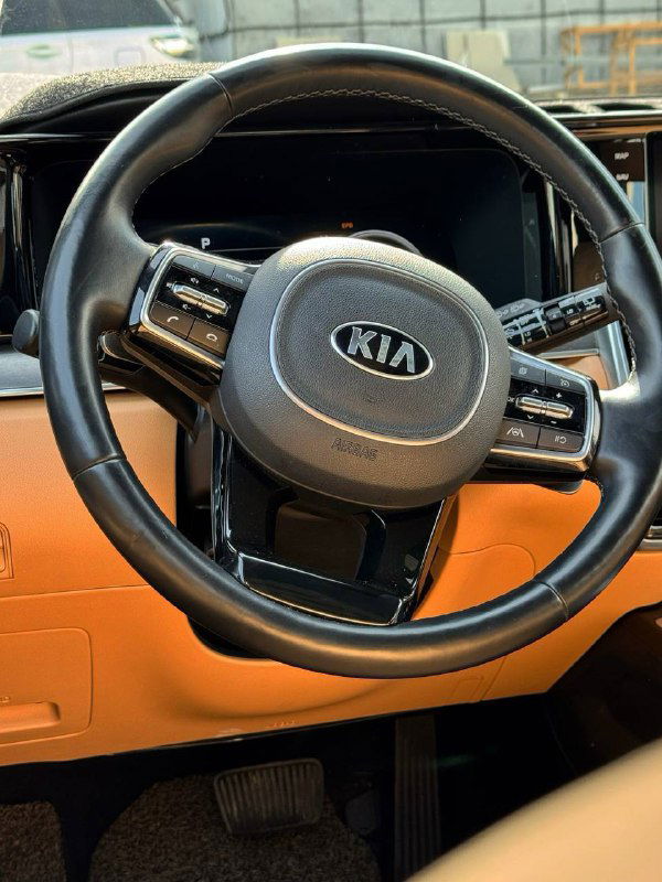 Крупный план рулевого колеса KIA с логотипом; чёрная отделка руля, элементы управления и часть оранжевой кожаной торпеды видны отчётливо.