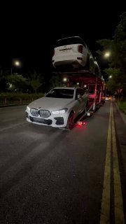 Автовоз с загруженными автомобилями на ночной городской улице в Пусане, вид спереди на BMW на нижней палубе и авто сверху