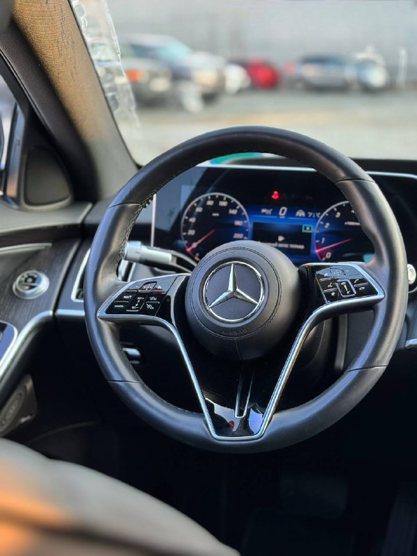 Руль и приборная панель Mercedes S 350d: крупный план рулевого колеса с эмблемой Mercedes и цифровой приборной панелью на фоне стоянки.