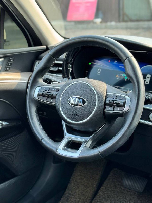 Крупный план руля KIA внутри салона K5, видны кнопки управления на руле и цифровая приборная панель за ним.