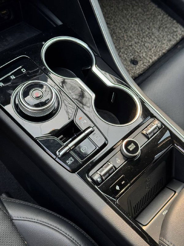 Крупный план центральной консоли KIA K5: шайба переключения передач, подстаканники и кнопки управления в глянцевой вставке.