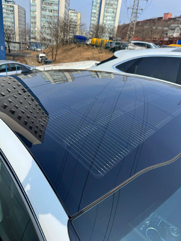 Крупный план стеклянной крыши KIA K5 с интегрированной солнечной панелью, видны фотоэлементы и отражение зданий на панели.