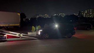 Ночной кадр: автомобиль съезжает с грузовой рампы возле автовоза, на фоне огней города и жилых домов, подготовка к отправке в порт.