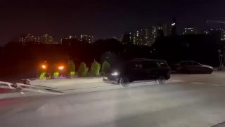 Ночной кадр приближения автомобиля к рампе погрузки, видны фары и городской горизонт на заднем плане, операция в Пусане.