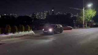 Крупный ночной кадр автомобиля с включёнными фарами на парковке у погрузочной зоны, вдалеке видна городская застройка и огни.