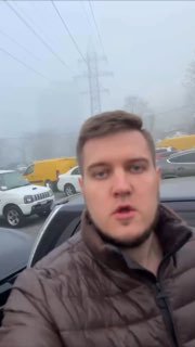 Вертикальное селфи мужчины на стоянке автомобилей: на заднем плане несколько машин и желтый фургон, туман и опоры ЛЭП.