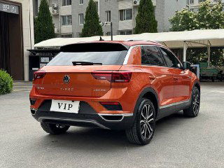 Вид сзади на оранжевый Volkswagen T‑Roc — задние фонари, эмблема VW на багажнике и оформленный бампер, автомобиль на городской площадке.