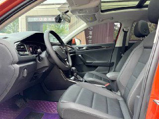 Передние сиденья Volkswagen T‑Roc — чёрная обивка, центральный тоннель, элементы управления и панорамная крыша над салоном, видно пространство для водителя и пассажира.