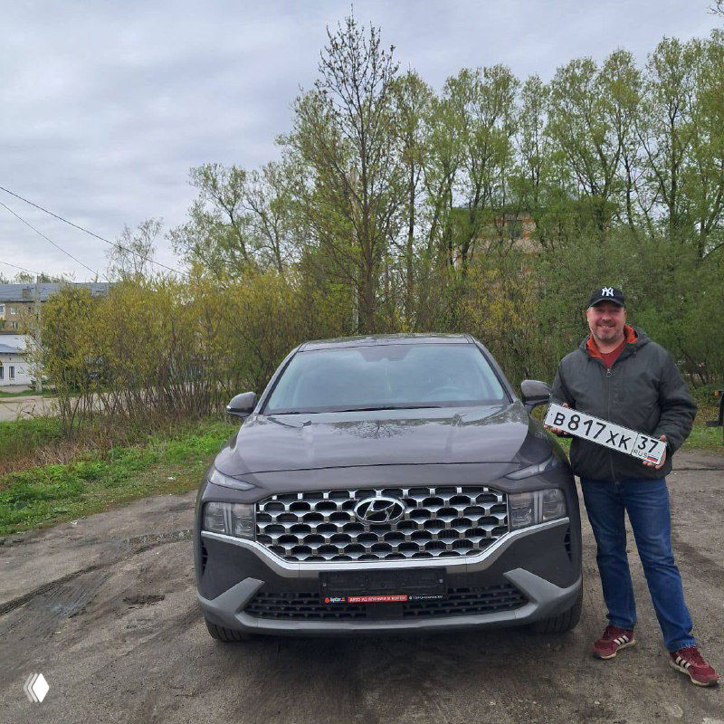 Вид спереди Hyundai Santa Fe: крупная решётка радиатора, эмблема и фары на фоне зелёных деревьев; автомобиль доставлен из Южной Кореи