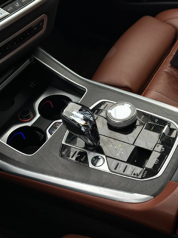 Блок селектора передач и управляющий джойстик BMW X7 на центральной панели — хромированные элементы и отделка под дерево рядом с кожаными креслами.