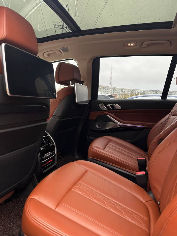 Вид на второй ряд сидений BMW X7 в коричневой коже: раскладные мониторы, простор для пассажиров и панорамная крыша на заднем плане.