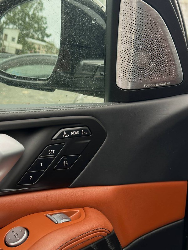 Деталь дверной панели BMW X7: элементы управления сиденьями, динамик мультимедийной системы и коричневые кожаные вставки в интерьере.