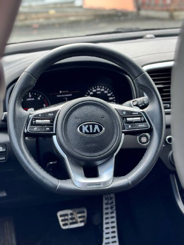 Фотография рулевого колеса KIA Sportage с логотипом KIA; видна приборная панель, мультикнопки на руле и часть торпеды — интерьер передней части салона