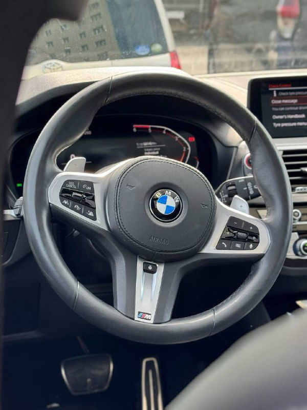 Крупный план рулевого колеса BMW с логотипом, элементы управления и часть цифровой приборной панели на фоне