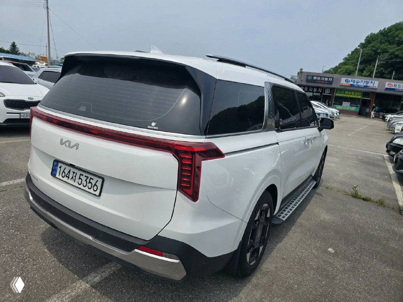 KIA Carnival 2024 — вид сзади и боковой проём, белый кузов, задняя светотехника и номерной знак, авто на стоянке в Корее.