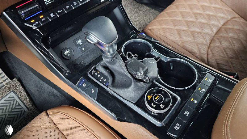 Центральный тоннель KIA Mohave 2022 — селектор коробки передач, регуляторы привода и подстаканники в интерьере с коричневой отделкой.