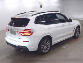 Задняя часть белого BMW X3 2020 на паркинге — вид сзади с включёнными задними фарами и заметными выхлопными патрубками.