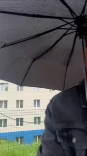 Фото стоянки в дождливый день: раскрытый тёмный зонт и часть куртки на фоне фасада дома и припаркованных автомобилей.