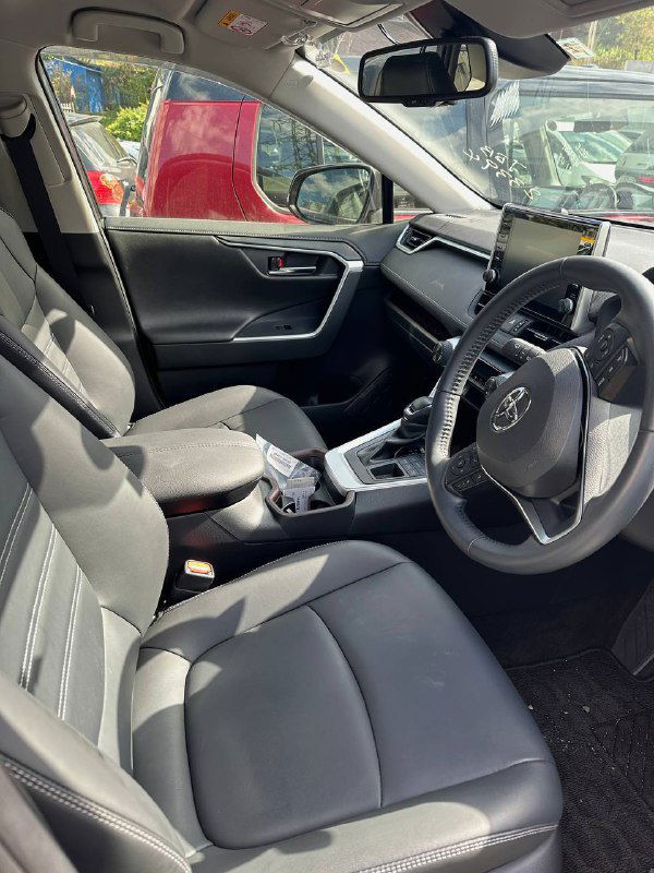 Снимок салона Toyota RAV4 2021: передние сиденья, руль, центральная консоль и мультимедийный экран, тканевая отделка, общий ракурс из дверного проёма.