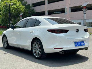Белая Mazda 3 2021, вид сзади справа на стоянке, показаны задние фонари и форма багажника, фото для оценки кузова при импорте.