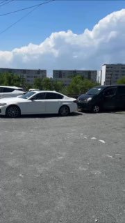 Автомобильная стоянка с несколькими легковыми машинами на асфальтовой площадке, на фоне жилых домов и чистого голубого неба, общий вид.