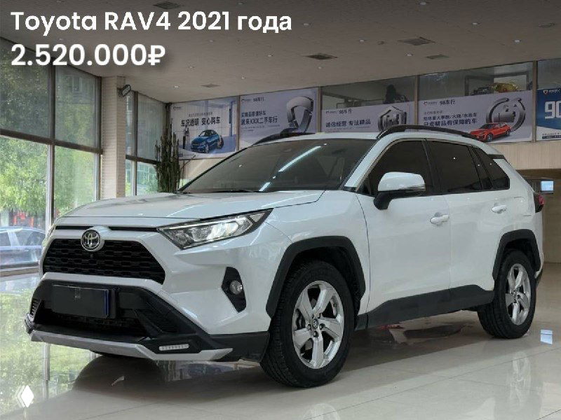 Фото белого Toyota RAV4 2021 года спереди в дилерском зале; вид передней части, решетка, фары и линия кузова автомобиля.
