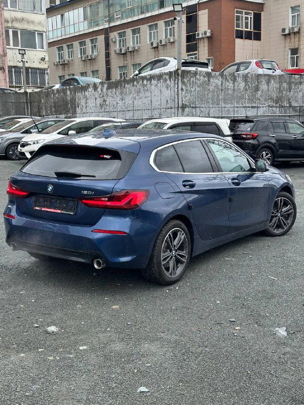 Синий BMW 120i 2022 года, вид сзади и в профиль, открытая задняя часть кузова, автомобиль припаркован у стены с зданиями.