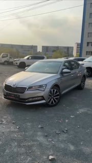 Серебристый Skoda Superb, припаркованный на стоянке перед зданием, вид спереди-сбоку — иллюстрация к видеообзору и описанию комплектации автомобиля.
