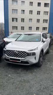 Белый Hyundai Santa Fe на парковке у многоэтажного дома; кадр показывает переднюю часть и бок, демонстрирует внешний вид и состояние кузова.