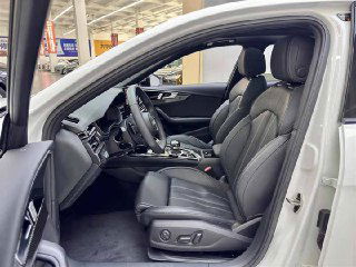 Передние сиденья Audi A4L 2022: вид со стороны открытой двери, чёрная кожаная обивка, электроприводы регулировки и просторная кабина на фоне склада автомобилей.