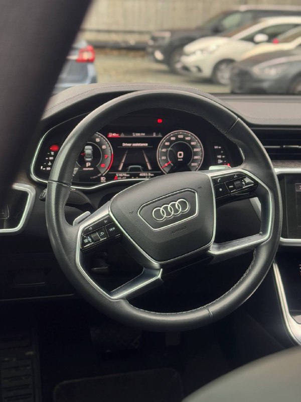 Крупный план рулевого колеса Audi A6 с логотипом, элементы управления на колесе и цифровая приборная панель с информацией о состоянии автомобиля.