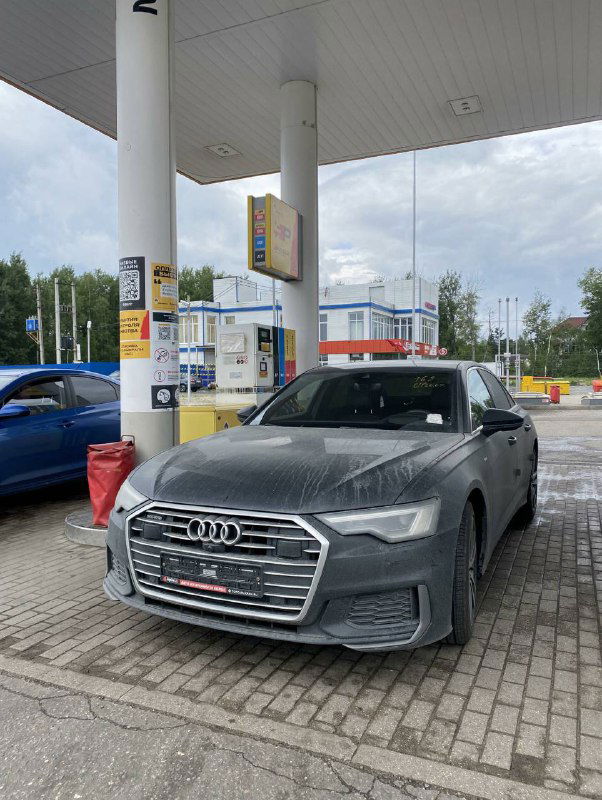 Фронтальный план Audi A6 на АЗС: видны решётка радиатора, фары и передняя часть кузова автомобиля клиента, облачная погода и заправочная инфраструктура.