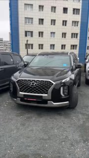 Чёрный Hyundai Palisade на парковке у многоэтажного жилого дома — фронтальный план автомобиля, вид спереди и справа, для видеообзора.