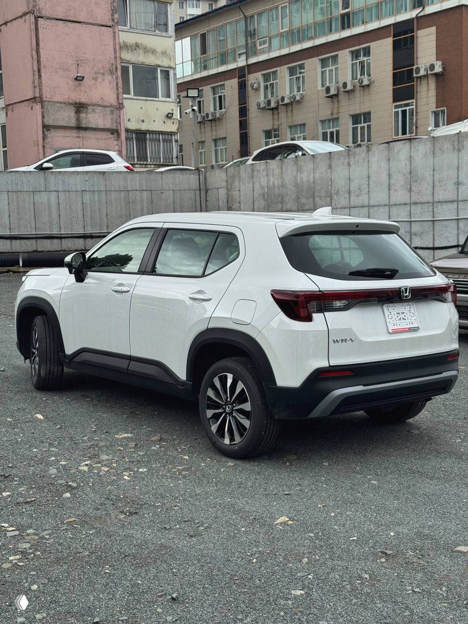 Вид сзади и сбоку на белый Honda WR‑V: кузов, задние фонари и колёсные диски на гравийной площадке у стоянки.