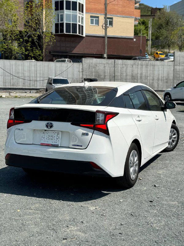 Toyota Prius 2022 белого цвета — вид сзади: багажник, задние фонари и бампер на фоне дворовой ограды и зданий.