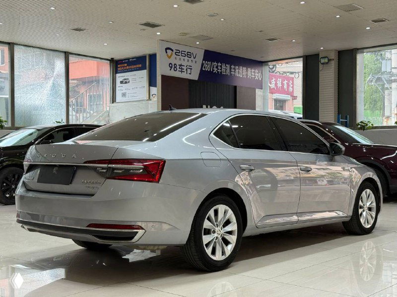 Задний план Skoda Superb в выставочном зале: серебристое купеобразное седан-окраска, вид сзади и рефлексы витрин на кузове.