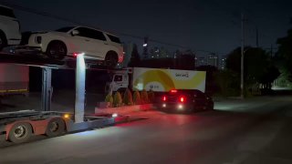 Вечерняя сцена: частичный вид автовоза и задние габаритные огни, на прицепе видны белые кроссоверы и городские огни порта Пусан на заднем плане.