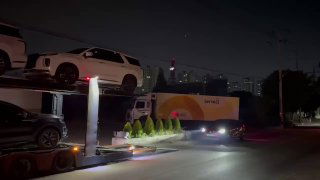 Крупный план: прицеп автовоза с белыми автомобилями и подсветкой, вид сбоку ночью, фон — складские и городские здания порта Пусан.