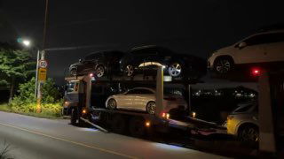 Ночной общий план: автовоз загружен разными моделями — внедорожники и седаны — движется по дороге у зелёной придорожной зоны рядом с портом Пусан.