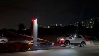 Ночной кадр: белый компактный кроссовер на нижней палубе автовоза, движение по освещённой дороге, городской силуэт и портовые огни на фоне.