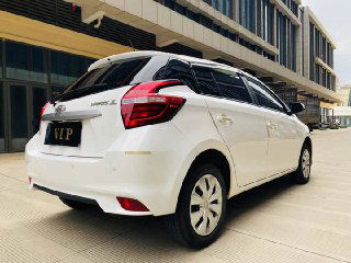 Белая Toyota Yaris L, задняя часть автомобиля на фоне городской застройки; вид демонстрирует состояние кузова и фонари.