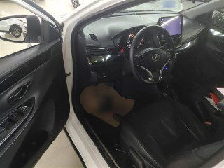 Вид с открытой водительской двери на передний ряд Toyota Yaris L 2022: кожаные сиденья, рулевая колонка и чистый интерьер для оценки общего состояния.