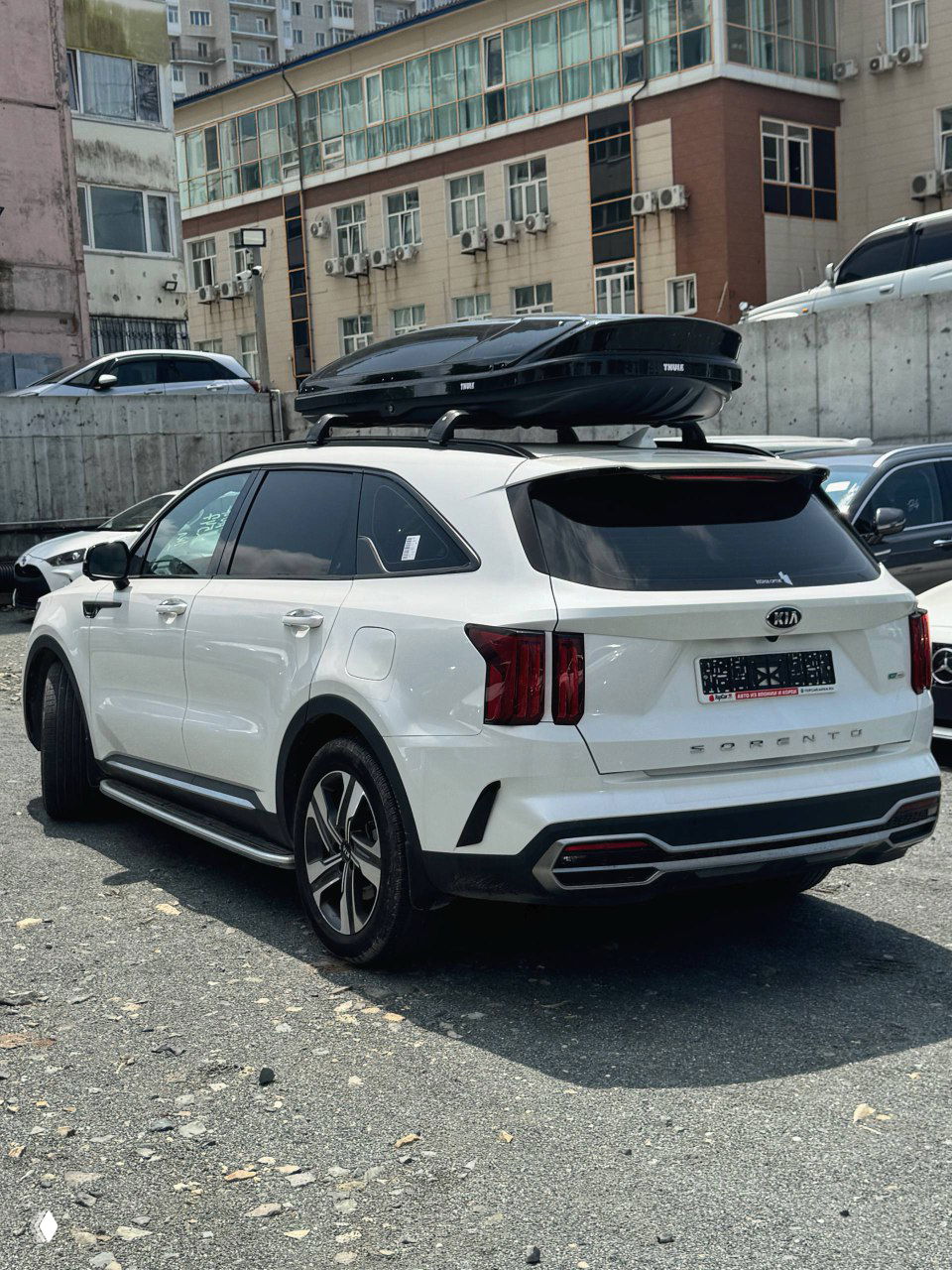 KIA Sorento 2021 вид сзади/сбоку в белом цвете с багажным боксом на крыше, видна эмблема и задняя оптика автомобиля.