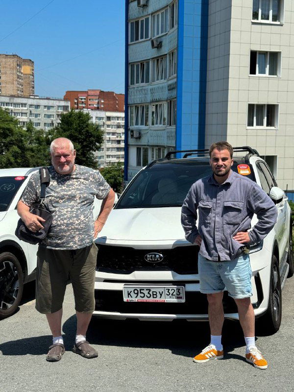 Клиент Сергей и сотрудник компании стоят рядом с белым Kia Sorento во дворе офиса TopCar во Владивостоке: передача автомобиля клиенту после оформления.