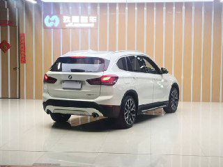 BMW X1 xDrive 25Li 2021 — вид сзади и под углом, белый кузов, задние фонари и бампер в выставочном пространстве на фоне декоративной стенки.