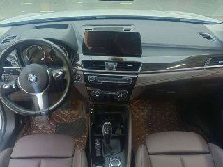 Интерьер BMW X1 xDrive 25Li 2021 — панорамный вид на переднюю панель, дисплей мультимедиа, руль и передние сиденья в коричневой обивке.