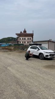 Белый кроссовер на гравийной площадке: сотрудник присел и фотографирует автомобиль перед отправкой, на фоне складского здания и пасмурного неба.