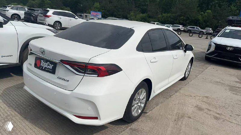 Toyota Corolla 2022, вид сзади слева; белый кузов, задние фонари и багажник видны отчётливо, автомобиль размещён среди других машин на площадке поставщика.